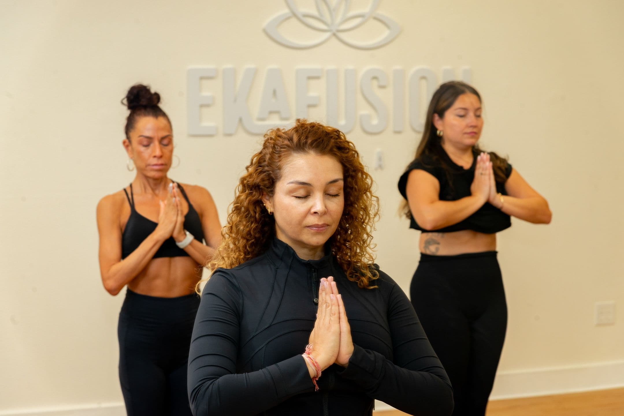 Three women practice yoga with hands at heart inside Ekafusion Wellness.