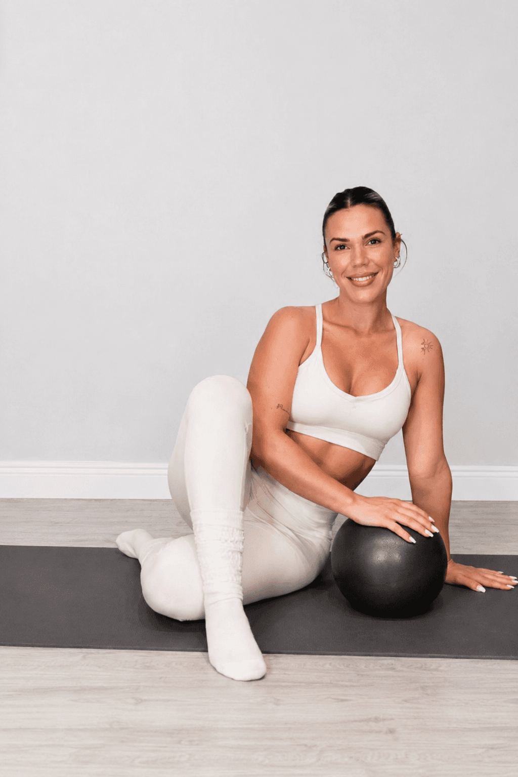 Caro sits on a Pilates mat beside a black exercise ball.